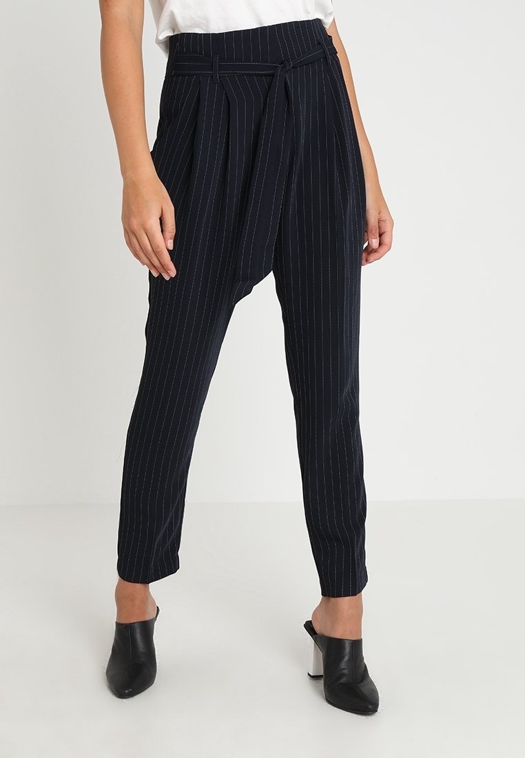 Pantalons bleu marine à rayures fines avec une coupe fuselée, dotés d'une taille haute, de passants de ceinture et de plis à l'avant. Associés à des mules noires à talons.