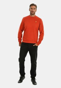 Roter Rundhalsausschnitt-Pullover mit gerippten Bündchen und Saum, kombiniert mit schwarzen Jeans und schwarzen Turnschuhen. Mit gesticktem Logo auf der Brust.