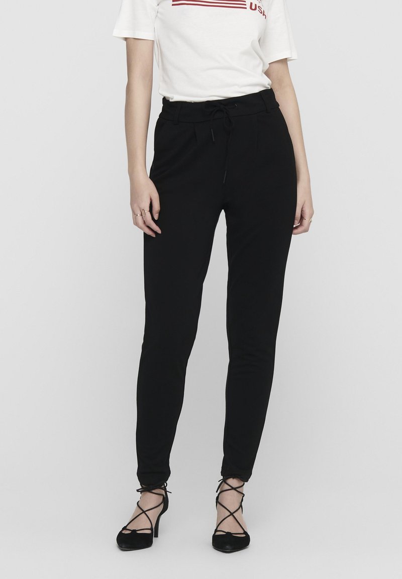 Femme portant un pantalon noir taille haute slim avec un cordon de serrage et des talons aiguilles noirs à lacets sur un fond uni.