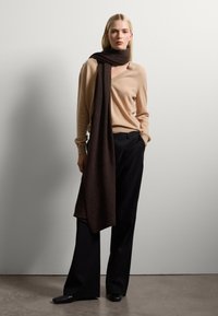 Jersey color beige con escote en V, combinado con una bufanda marrón oscuro y pantalones de pierna ancha negros. La bufanda cae sobre el hombro, con textura suave.