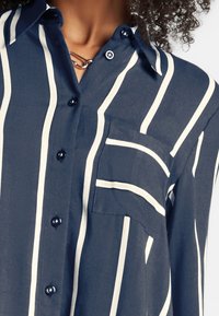 Primer plano de una persona que lleva una camisa azul marino con rayas blancas verticales y un bolsillo en el pecho, parcialmente desabrochada para revelar una cadena de plata.