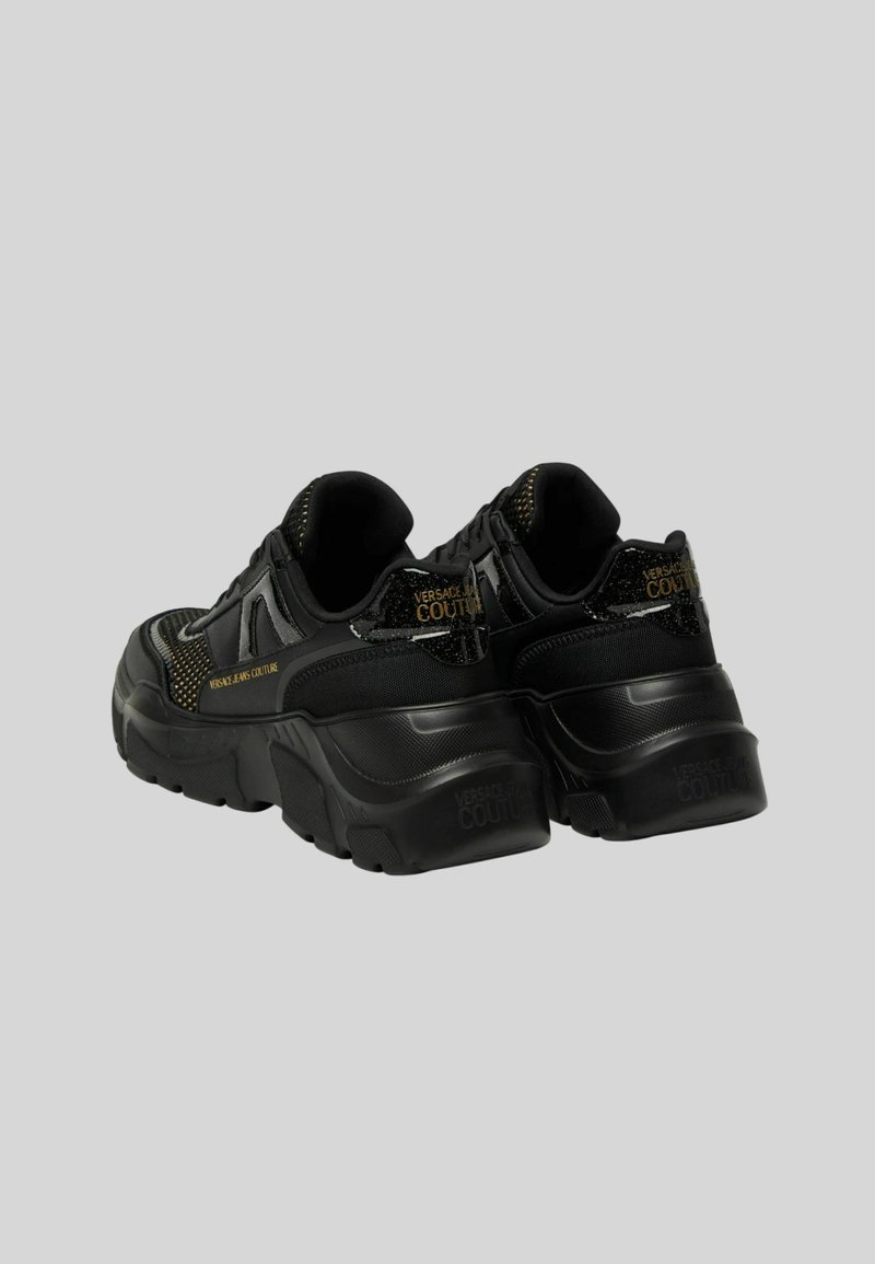Baskets noires avec une tige en mesh texturé, une semelle épaisse et des accents dorés, portant la marque "VERSACE COUTURE" au niveau du talon.