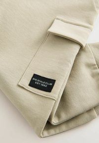 Beige sweatshirt med manchetter på ærmerne, udstyret med et sort mærke, der står "BIG SMILE CLUB EST.1982." Glat tekstur og enkelt design.