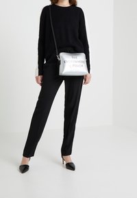 Pochette synthétique argentée avec le texte imprimé "THE EVERYWHERE POUCH", associée à un pull noir à côtes et un pantalon noir, portée avec des talons pointus.
