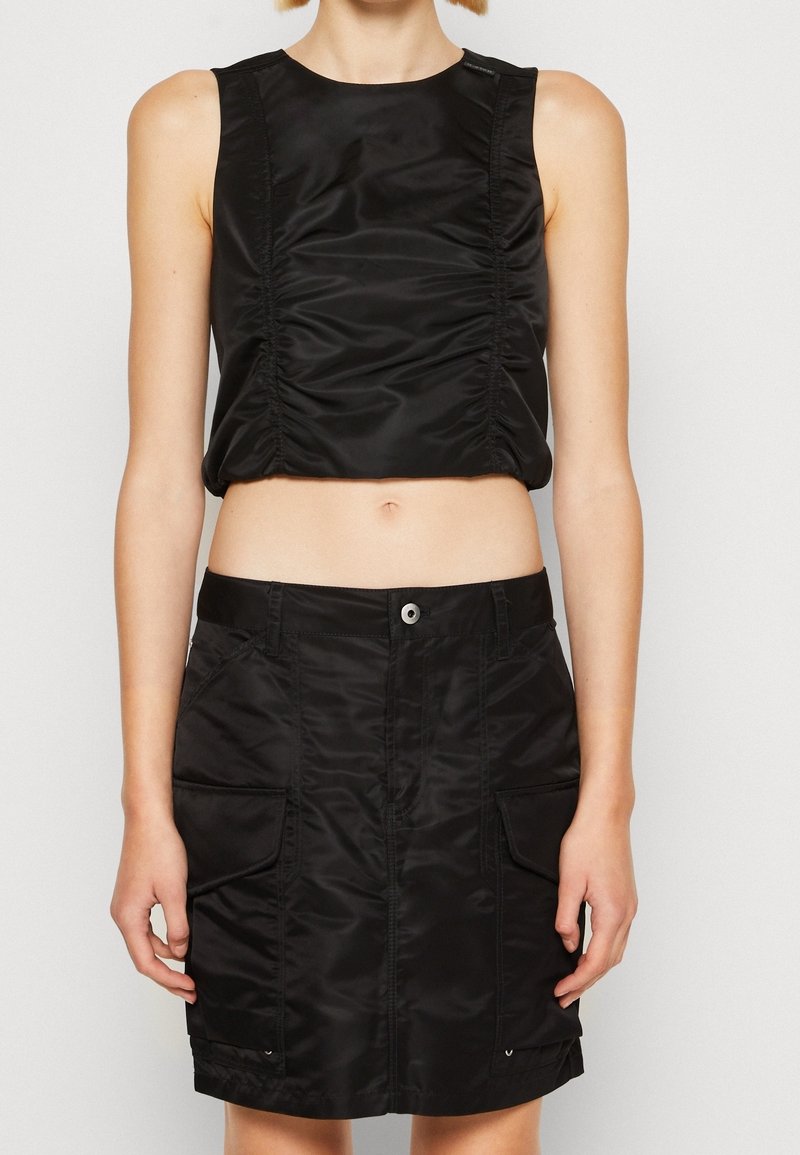 Femme portant un top court sans manches noir et une jupe noire taille haute avec poches avant et fermeture par boutons.