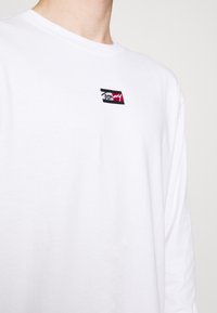 Person in einem weißen Langarmhemd mit einem kleinen rechteckigen schwarzen, weißen und roten Logo auf der oberen Brust.