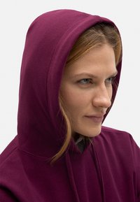 Sudadera burdeos con un tejido texturizado, que presenta una capucha con cordón y un ajuste relajado, que cubre parcialmente la cara de una persona.