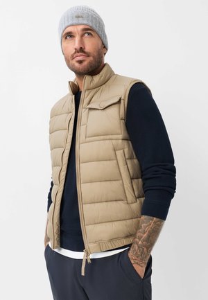 Mann mit Bart, der eine graue Mütze, eine beige gesteppte Weste, einen dunkelblauen Pullover und dunkle Hose trägt, steht mit den Händen in den Taschen vor schlichtem Hintergrund.