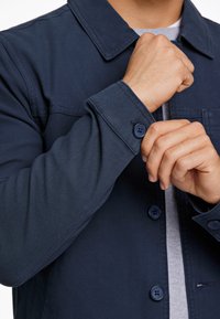 Donkerblauwe button-up jas met kraag, voorzien van een borstzak en knoopsluitingen bij de manchetten. Glad materiaal met zichtbare stikseldetails.