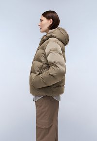 Donna in una giacca a vento beige con cappuccio, mani nelle tasche, che indossa pantaloni cargo marroni, mostrata di profilo contro uno sfondo chiaro e uniforme.
