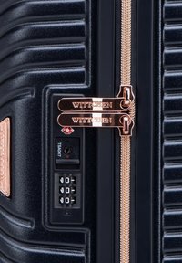 Valigia rigida blu navy con strisce in rilievo, hardware in oro rosato, doppie cerniere e lucchetto approvato TSA. Mostra il nome del marchio "WITTCHEN."