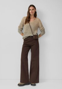 Vrouw in een beige gebreide trui, donkere bruine wijde broek, beige chunky laarzen en een kleine bruine schoudertas, staand met de handen achter zich.