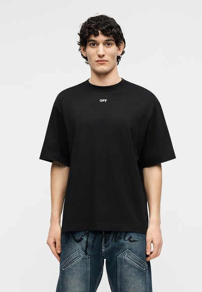 Νεαρός με σκούρα σγουρά μαλλιά, φορώντας μαύρο υπερμεγέθες t-shirt με την επιγραφή "OFF" και μπλε τζιν με ραμμένες λεπτομέρειες στην τσέπη.