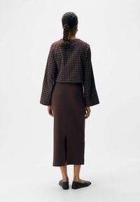 Vrouw met gevlochten haar, gekleed in een geruite lange mouwen top en een donkerbruine midi-rok met een split aan de achterkant, staande met haar rug naar de kijker.