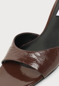 Gros plan d'une sandale ouverte en cuir verni marron avec une large bride avant et une doublure intérieure noire.