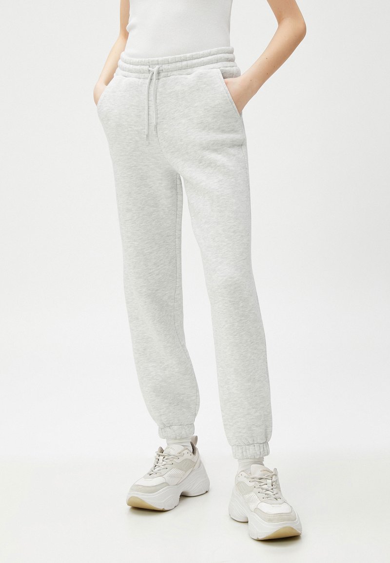 Persona con pantalones jogger de color gris claro con puños elásticos, zapatillas blancas gruesas y una camiseta sin mangas blanca, de pie con las manos en los bolsillos.