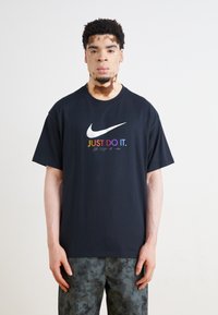 Juodas medvilninis marškinėliai su spalvingu "JUST DO IT." logotipu ir swoosh grafika. Trumpomis rankovėmis, laisvas prigludimas, tekstūruotas audinys.
