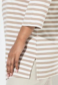 Pasiasta tunika z beżowej i białej bawełny, charakteryzująca się luźnym krojem, krótkimi rękawami i rozcięciami po bokach dla swobody ruchów.