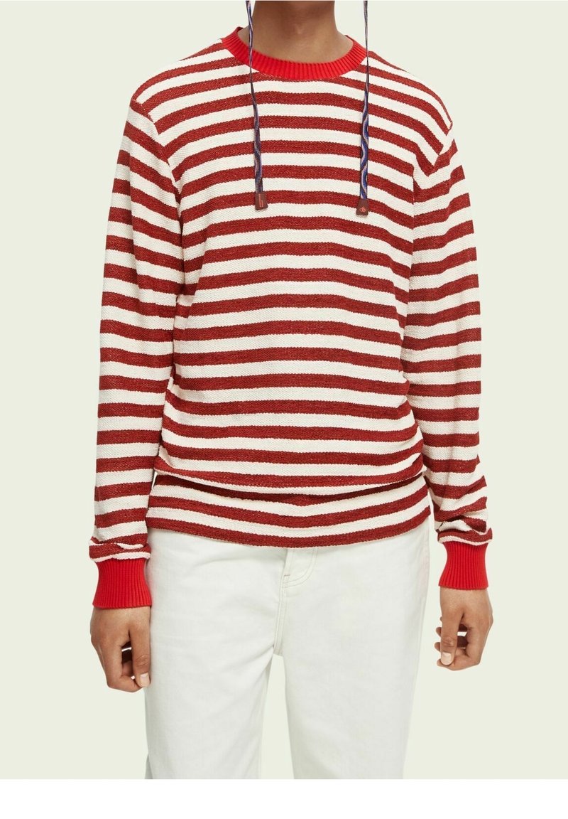 Personne portant un haut à manches longues rayé horizontalement rouge et blanc avec des poignets rouges et un pantalon blanc, sur un fond neutre.