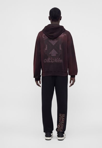 Homme portant un sweat à capuche rouge foncé avec un motif et le texte "A.C. Milan" dans le dos, un pantalon noir avec des logos de croix rouges sur la jambe inférieure, et des baskets noires.