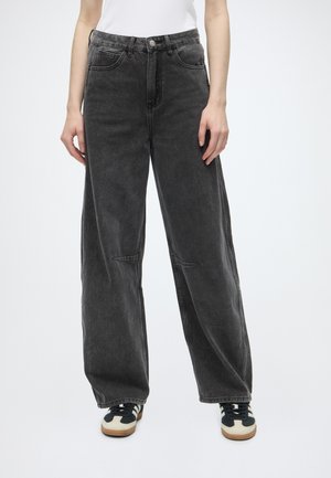 Person trägt locker sitzende schwarze gewaschene Jeans und weiße Sneakers mit schwarzen Schnürsenkeln, steht vor einem schlichten weißen Hintergrund.