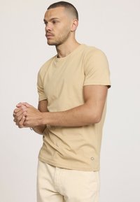 T-shirt en coton beige à manches courtes et col rond. Présente un petit accent en forme de diamant près de l'ourlet. Texture lisse et design ajusté.