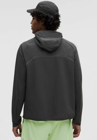 Man som bär en mörkgrå huva jacka och matchande mössa, sedd från ryggen, med ljusgröna shorts mot en enfärgad bakgrund.