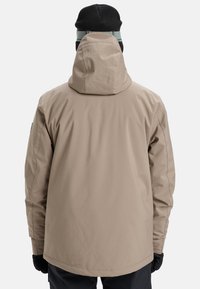 Beige waterproof jacket with a drawstring hood, long sleeves, and a straight cut. Textured fabric with minimal seams, worn with black gloves.