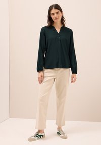 Dunkelgrüne Bluse mit einem V-Ausschnitt, langen Ärmel und Knopfleiste. Kombiniert mit beigen, weit geschnittenen Hosen und grauen Sneakers mit grünen Akzenten.