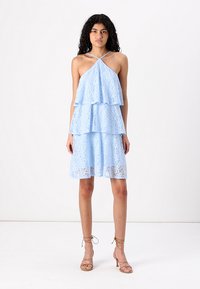 Svetlomodré čipkované šaty s vrstvenými volánmi a halterneckým výstrihom. Tkanina obsahuje kvetinové vzory, doplnené o sandále s pásikmi v nude farbe.