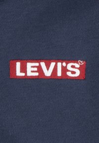 Námořnická modrá látka s červenou vyšitou obdélníkovou nášivkou s bílým textem "LEVI'S" a malým ochranným symbolem.