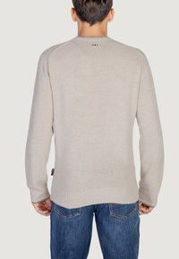 Hellbeige Pullover mit gerippten Bündchen und Saum, ausgestattet mit einem Rundhalsausschnitt und dezentem Branding auf der Rückseite. Getragen mit blauer Jeans.