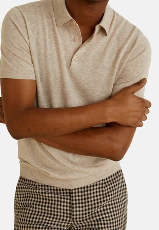 Homme portant un polo beige à manches courtes et un pantalon à carreaux noir et blanc, les bras croisés sur la poitrine.