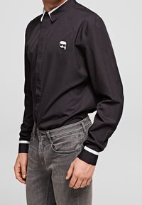 Černá košile na knoflíky s bílým límcem a manžetami, s vyšitým logem na hrudi. Tkanina se zdá hladká a odolná.