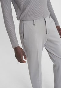 Pantaloni grigi chiari su misura con una finitura testurizzata, dotati di tasche laterali, zip sul davanti e passanti per cintura minimalisti. Top a maniche lunghe con righe visibile.