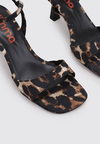 Sandalias de tacón alto en negro y marrón con un acabado suave, punta abierta y una delgada correa en el tobillo. Diseño minimalista con un tacón cuadrado.