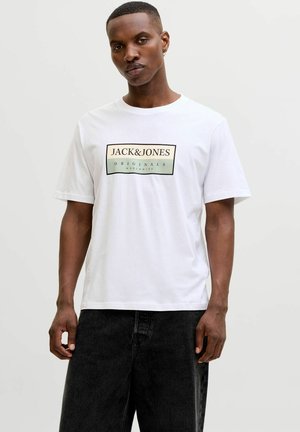 Mann mit kurzen Haaren, der ein weißes T-Shirt mit dem Aufdruck „JACK&JONES ORIGINALS WORLDWIDE“ und schwarze Hosen vor hellem einfarbigem Hintergrund trägt.