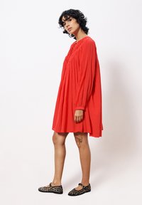 Rotes, langärmliges Kleid mit gerafftem Dekolleté und lockerer Passform. Getragen mit flachen Schuhen im Leopardenmuster und sichtbarem Tattoo am Bein.