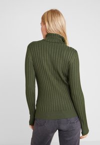 Femme portant un pull à col roulé côtelé vert olive et un jean gris foncé, tournée de dos sur un fond uni gris clair.