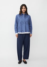 Niebieska koszula z guzikiem, ozdobiona wzorzystym haftem, w połączeniu z granatowymi spodniami o szerokich nogawkach i czarnymi butami, z białą koszulą pod spodem.