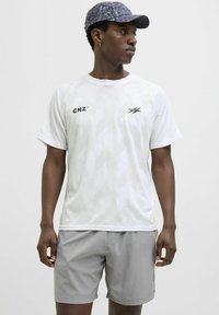 Hvid kortærmet sports t-shirt med geometrisk mønster, sort "CNZ" logo, parret med grå shorts. Modellen bærer en marineblå mønstret cap.