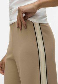 Beige atletische broek met een gladde textuur, voorzien van een zwart en crème verticale streep langs de zijkant. Hand op de taille geplaatst.