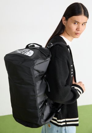 Persoon met lang haar, gekleed in een zwarte cardigan en lichte spijkerbroek, draagt een grote zwarte rugzak van The North Face.