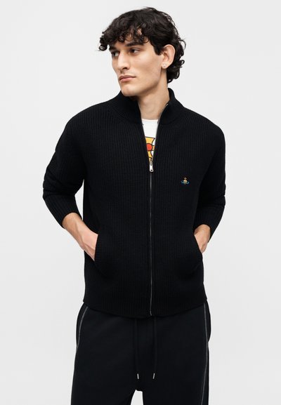 Μαύρο πλεκτό πουλόβερ με φερμουάρ και υψηλό γιακά, με δύο μπροστινές τσέπες και μικρό λογότυπο στο αριστερό στήθος. Φορεμένο πάνω από ένα λευκό T-shirt.