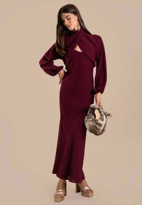 Bordeauxfarbenes Kleid mit langen Ärmeln, verdrehtem Ausschnitt und Ausschnittdetails; kombiniert mit einer metallicfarbigen Clutch und goldenen Plateauschuhen.