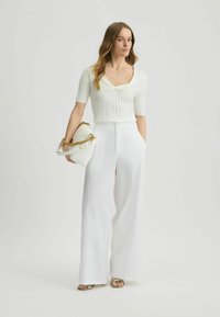 Weißes geripptes Oberteil mit quadratischem Ausschnitt, kombiniert mit weiten weißen Hosen. Hält eine weiße Tasche mit einer goldenen Kette. Texturierte Designelemente.