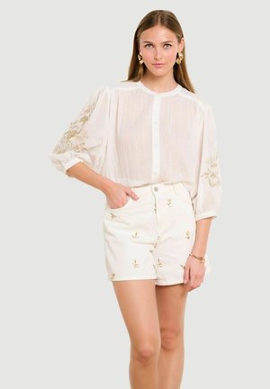 Žena v bielej priehľadnej blúzke s nafúknutými rukávmi a zlatou kvetinovou výšivkou, v kombinácii s bielymi vysokými šortkami s malými kvetinovými vzormi.