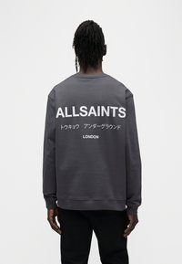 Γκρι φούτερ με μακριά μανίκια, με μεγάλο λευκό κείμενο στην πλάτη: "ALLSAINTS", ιαπωνικό κείμενο και "LONDON" σε μικρότερα γράμματα.