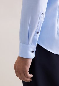 Manica di camicia azzurro chiaro con una texture liscia, dotata di bottoni scuri e di un dettaglio floreale ricamato vicino al polsino.
