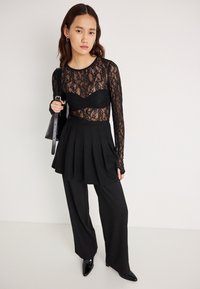 Μαύρο τοπ με μακριά μανίκια από δαντέλα, φορεμένο πάνω από μαύρο σουτιέν, συνδυασμένο με μαύρη φούστα μίνι με φτερό και ίσια μαύρη παντελόνα. Γυαλιστερά μαύρα τακούνια.